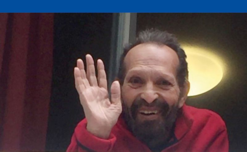 Ein älterer Mann winkt lächelnd und fröhlich aus einem Fenster.