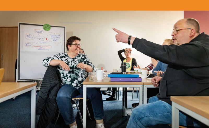 Mehrere Erwachsene sitzen in einem Unterrichts- oder Bildungskontext und wirken fröhlich.